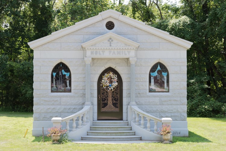 Private walk in family mausoleum made with granite stone has steps and railing with intricate details. Decorative detailing on the door and stained glass windows incorporate personal family touches
