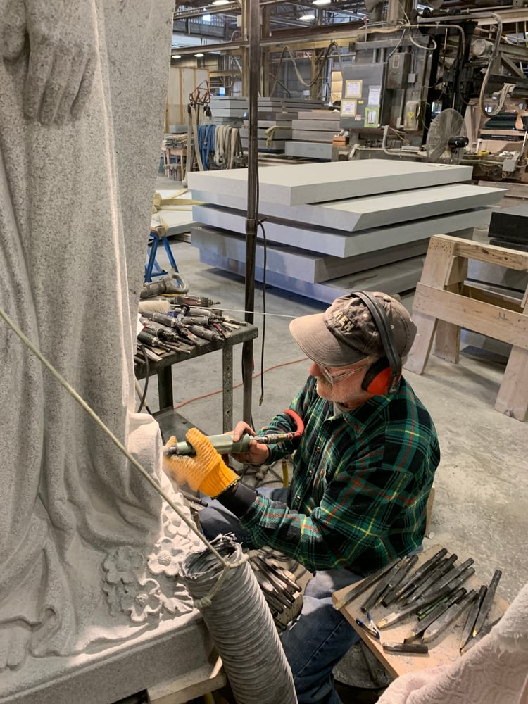 A Rock of Ages master stone carver uses tools to intricately craft details into a Bethel White granite sculpture on the Craftsmen Center Floor